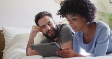 Video of happy diverse couple relaxing at home, lying on bed using tablet. Happiness, communication, inclusivity, free time, togetherness and domestic life.