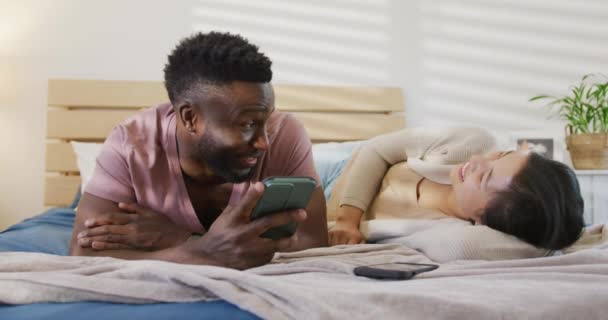 Happy Diverse Couple Using Smartphone Lying Bedroom Spending Quality Time — Vídeo de Stock