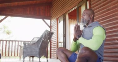 Happy senior african american man spending time in log cabin, doing yoga and meditating, slow motion. Free time, domestic life and nature concept.