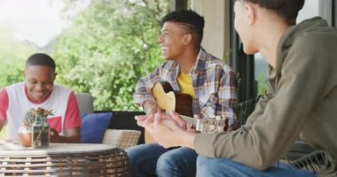 Happy diverse male teenage friends playing guitar at home, slow motion. Spending quality time at home together.