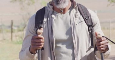 Portrait of happy senior african american man hiking with trekking poles on sunny day, slow motion. Hiking, active lifestyle and nature concept.