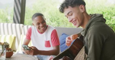 Happy diverse male teenage friends playing guitar at home, slow motion. Spending quality time at home together.