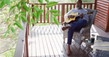 Happy senior african american man spending time in log cabin, playing guitar on balcony, slow motion. Free time, domestic life and nature concept.