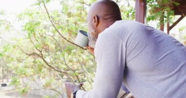 Happy senior african american man in log cabin, drinking coffee on balcony, slow motion. Free time, domestic life and nature concept.