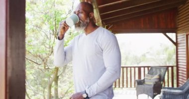 Happy senior african american man in log cabin, drinking coffee on balcony, slow motion. Free time, domestic life and nature concept.