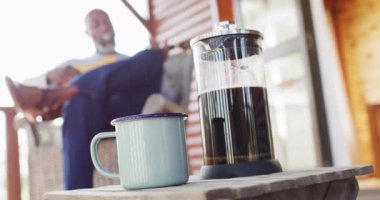 Mug of coffee over senior african american man in log cabin, playing guitar on balcony, slow motion. Free time, domestic life and nature concept.