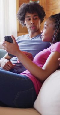 Kanepede oturan ve kulübede akıllı telefon kullanan Afro-Amerikan çiftin dikey videosu. Boş zaman ve ev hayatı kavramı.