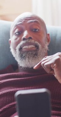 Vertical video of senior african american man spending time in log cabin using smartphone. Free time, domestic life and nature concept.