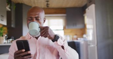 Happy senior african american man in log cabin, using smartphone and drinking coffee, slow motion. Free time, domestic life and communication concept.