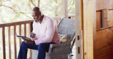 Happy senior african american man spending time in log cabin, using tablet on balcony, slow motion. Free time, domestic life and communication concept.
