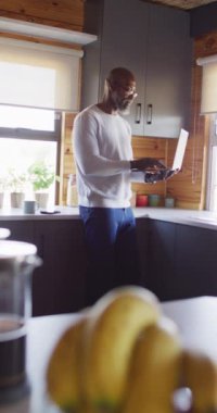 Vertical video of senior african american man spending time in log cabin using laptop. Free time, domestic life and nature concept.