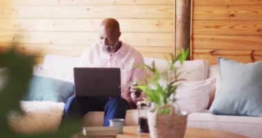 Happy senior african american man in log cabin, using laptop for online shopping, slow motion. Free time, domestic life and communication concept.