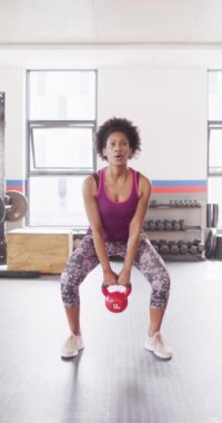 Beyzbol salonunda kettlebell sallayan kararlı Afrikalı Amerikalı kadının dikey videosu. Egzersiz, spor ve sağlıklı yaşam tarzı..