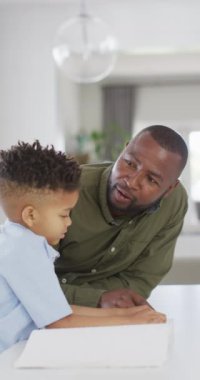 Vertical video of african american man and his son sitting at table and reading braille. Spending quality time at home.
