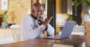 Happy senior african american man in log cabin, using laptop for video call, slow motion. Free time, domestic life and communication concept.