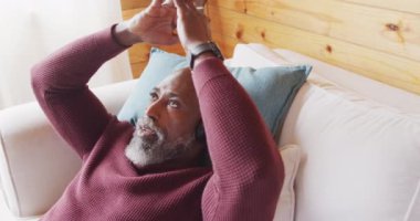 Happy senior african american man in log cabin, laying on sofa and using headphones, slow motion. Free time, domestic life and leisure concept.