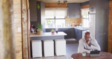 Senior african american man in log cabin, sitting at table and using tablet, slow motion. Free time, domestic life and communication concept.