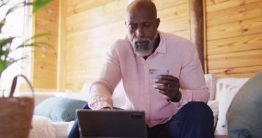 Happy senior african american man in log cabin, using tablet for online shopping, slow motion. Free time, domestic life and communication concept.