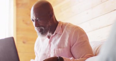 Happy senior african american man in log cabin, sitting on sofa and using laptop, slow motion. Free time, domestic life and communication concept.