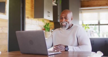 Happy senior african american man in log cabin, using laptop for video call, slow motion. Free time, domestic life and communication concept.
