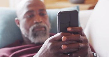 Happy senior african american man in log cabin, sitting on sofa and using smartphone, slow motion. Free time, domestic life and communication concept.