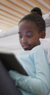 Evdeki ranzada oturup fotokopi çeken mutlu Afrikalı Amerikalı bir kızın dikey videosu. Teknoloji, iletişim ve ev hayatı.