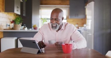 Happy senior african american man in log cabin, using tablet for online shopping, slow motion. Free time, domestic life and communication concept.