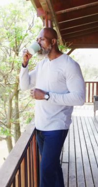 Vertical video of senior african american man spending time in log cabin drinking coffee. Free time, domestic life and nature concept.