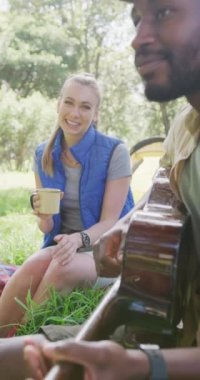 Vertical video of happy diverse couple camping, drinking tea and playing guitar in park, slow motion. Nature, hiking and park concept.