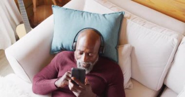 Happy senior african american man in log cabin, laying on sofa and using smartphone, slow motion. Free time, domestic life and leisure concept.