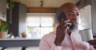 Happy senior african american man in log cabin, using smartphone and drinking coffee, slow motion. Free time, domestic life and communication concept.
