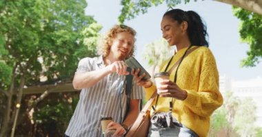 Happy diverse couple hailing taxi, using smartphone and holding takeaway coffee in city. City break, travel and hospitality concept.