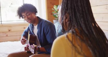 Mutlu Afro-Amerikan çift gitar çalıyor, odunlukta kanepede oturuyor, ağır çekimde. Yaşam tarzı, ev hayatı, kırsal ve doğa kavramı.