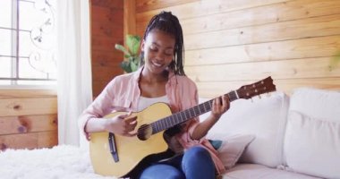 Mutlu Afro-Amerikan kadın kanepede oturuyor ve odunlukta gitar çalıyor, ağır çekimde. Yaşam tarzı, ev hayatı, kırsal ve doğa kavramı.