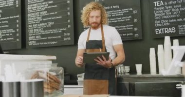 Portrait of happy caucasian male barista using tablet behind counter in cafe. Local business owner and hospitality concept.