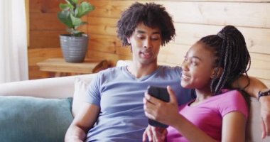 Akıllı telefon kullanan mutlu Afro-Amerikan çifti odunlukta koltukta ağır çekimde oturuyorlar. Ev hayatı, kırsal alan ve doğa kavramı.
