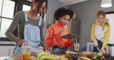 Diverse teenager girls friends cutting fruit and using tablet in kitchen, slow motion. Friends, leisure, spending quality time at home concept.