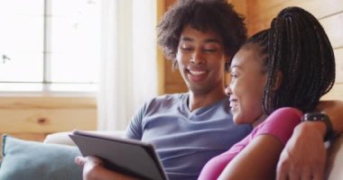 Mutlu Afro-Amerikan çift tablet kullanıyor, odunlukta kanepede oturuyor, ağır çekimde. Yaşam tarzı, ev hayatı, kırsal ve doğa kavramı.