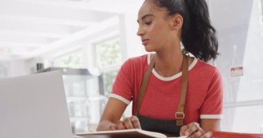 Biracial female barista wearing apron using laptop at table in her cafe. Local business owner and hospitality concept.
