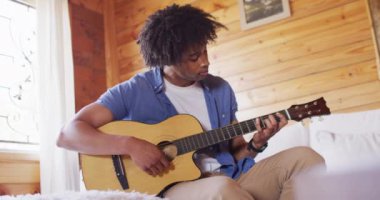 Mutlu Afro-Amerikan adam kanepede oturuyor ve odunlukta gitar çalıyor, ağır çekimde. Yaşam tarzı, ev hayatı, kırsal ve doğa kavramı.