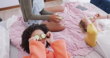 Yüzünde salatalık dilimleri, yavaş çekimde yatakta yatan farklı genç kızlar. Arkadaşlar, eğlence, ev konseptinde kaliteli zaman geçirme..