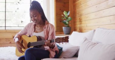 Mutlu Afro-Amerikan kadın kanepede oturuyor ve odunlukta gitar çalıyor, ağır çekimde. Yaşam tarzı, ev hayatı, kırsal ve doğa kavramı.
