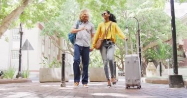 Happy diverse couple walking with luggage on sunny day in city. City break, travel and hospitality concept.