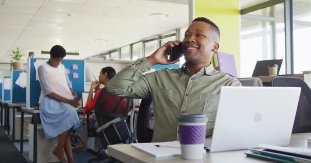 アフリカ系アメリカ人のビジネスマンがノートパソコンを使ってオフィスでスマートフォンで話をしている オフィスで働くこと ビジネスコンセプト — ストック動画