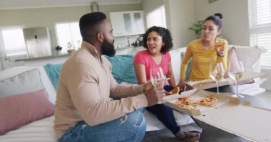 Three diverse male and female friends eating pizza and having wine in living room in slow motion. Friendship, leisure and lifestyle concept.
