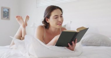 Happy caucasian woman relaxing, lying in bed and reading book. Spending quality time at home concept.