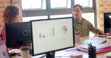 Happy caucasian creative talking with diverse colleagues at desk in casual office, slow motion. Start up business, teamwork and communication concept.