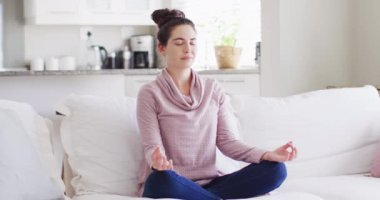 Happy caucasian woman sitting on couch and meditating in living room. Spending quality time at home concept.