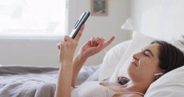 Kafkasyalı mutlu bir kadın yatakta uzanıyor ve akıllı telefon kullanıyor. Ev kavramında kaliteli zaman geçirmek.