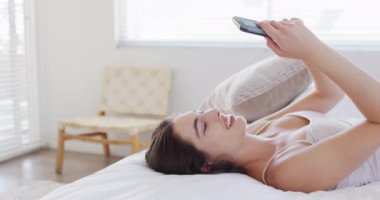 Kafkasyalı mutlu bir kadın yatakta uzanıyor ve akıllı telefon kullanıyor. Ev kavramında kaliteli zaman geçirmek.